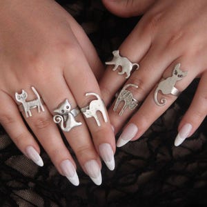 May include: A collection of six silver-toned metal rings, each featuring a distinct cat-shaped design. The rings are worn across the fingers, showcasing various feline silhouettes including sitting, stretching, and walking poses in a metallic finish.