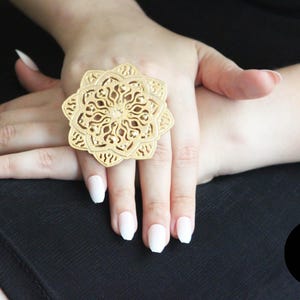 May include: A gold-toned ring with a large, intricate, circular design. The ring is worn on the index finger of a hand with white nail polish.