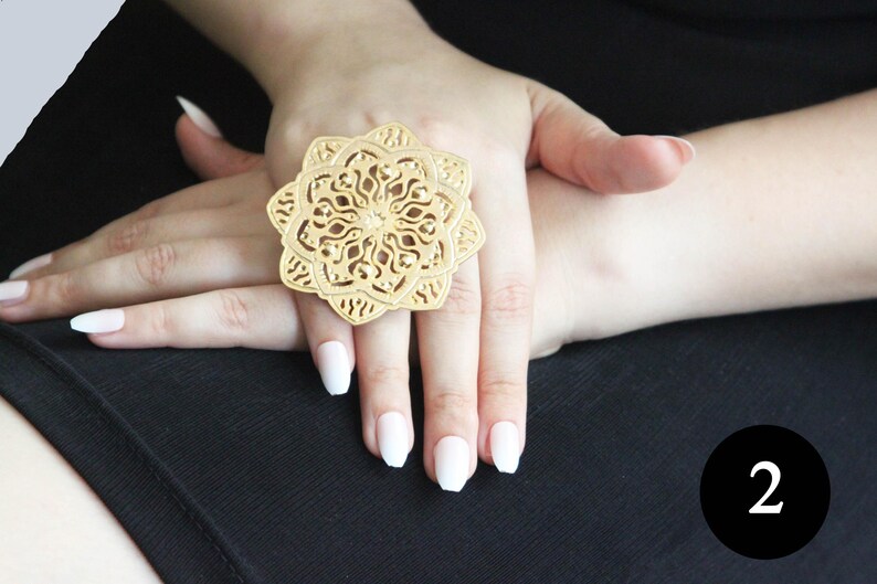 May include: A gold-toned ring with a large, intricate, circular design. The ring is worn on the index finger of a hand with white nail polish.