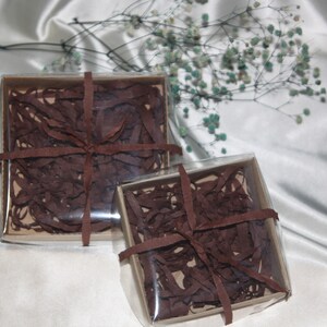 May include: Two clear gift boxes with brown shredded paper and brown ribbon. The boxes are on a white satin background.