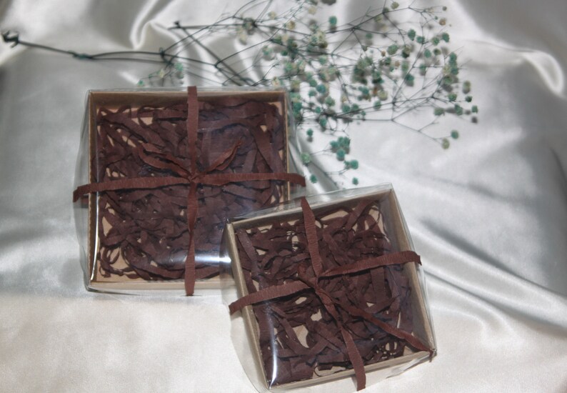 May include: Two clear gift boxes with brown shredded paper and brown ribbon. The boxes are on a white satin background.