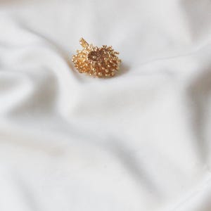 May include: A gold-coloured, textured, round pendant with a delicate design. The pendant is on a white fabric background.