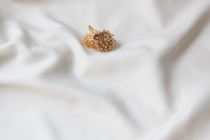 May include: A gold-coloured, textured, round pendant with a delicate design. The pendant is on a white fabric background.