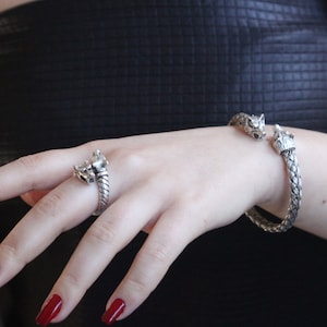 May include: A silver bracelet and ring featuring wolf head designs. The bracelet is a braided design with wolf heads at each end. The ring is a similar design with a wolf head on the top.