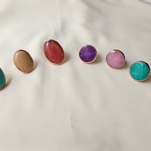 May include: Seven oval and round rings with different colored stones. The stones are turquoise, wood, red, purple, pink, green, and blue.