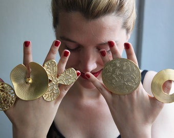 May include: A woman's hands wearing five gold rings. The rings are all different shapes and sizes, and some have intricate designs. The rings are made of a shiny gold metal.