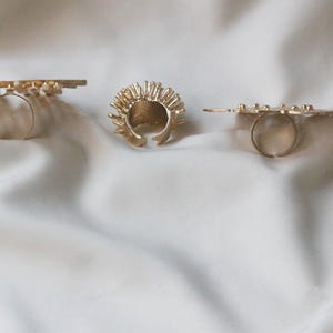May include: Three gold rings with different designs. The first ring is a thin band with a geometric pattern. The second ring is a thick band with a spiky design. The third ring is a thin band with a geometric pattern.
