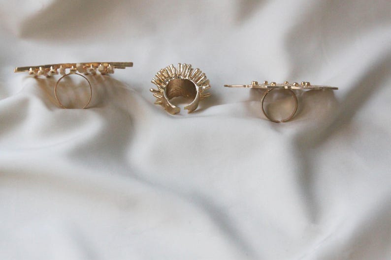 May include: Three gold rings with different designs. The first ring is a thin band with a geometric pattern. The second ring is a thick band with a spiky design. The third ring is a thin band with a geometric pattern.