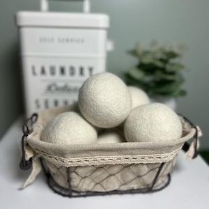 May include: A white wire basket with a beige fabric liner and lace trim holds a group of six white wool dryer balls. The basket is sitting on a white surface.