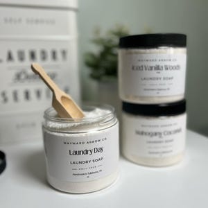 May include: Three white jars of laundry soap with black lids. The front jar is labelled "Wayward Arrow Co. Laundry Day Laundry Soap since 2020 Handmade in Tullahoma, TN". The middle jar is labelled "Iced Vanilla Woods Laundry Soap". The back jar is labelled "Refreshing Coconut Laundry Soap". A wooden scoop is next to the front jar.