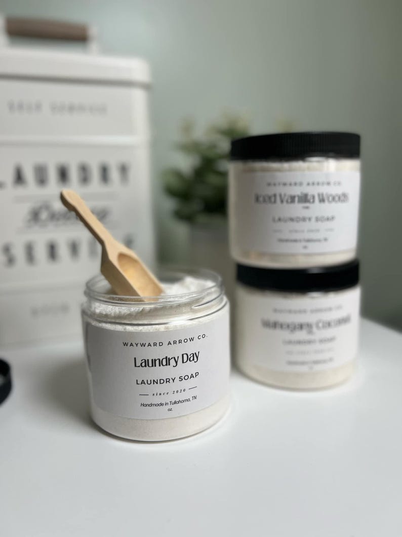May include: A collection of laundry products, including jars of laundry soap and a wooden scoop. The jars have black lids and white labels with text. One jar is open, revealing the soap inside. The text on the labels includes "Laundry Day" and "Iced Vanilla Woods."