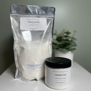 May include: A clear plastic bag and a white jar of laundry soap sit on a white surface. The bag is labeled "Sweet Grace Laundry Soap" and the jar is labeled "Laundry Day Laundry Soap". A small plant in a white pot is in the background.