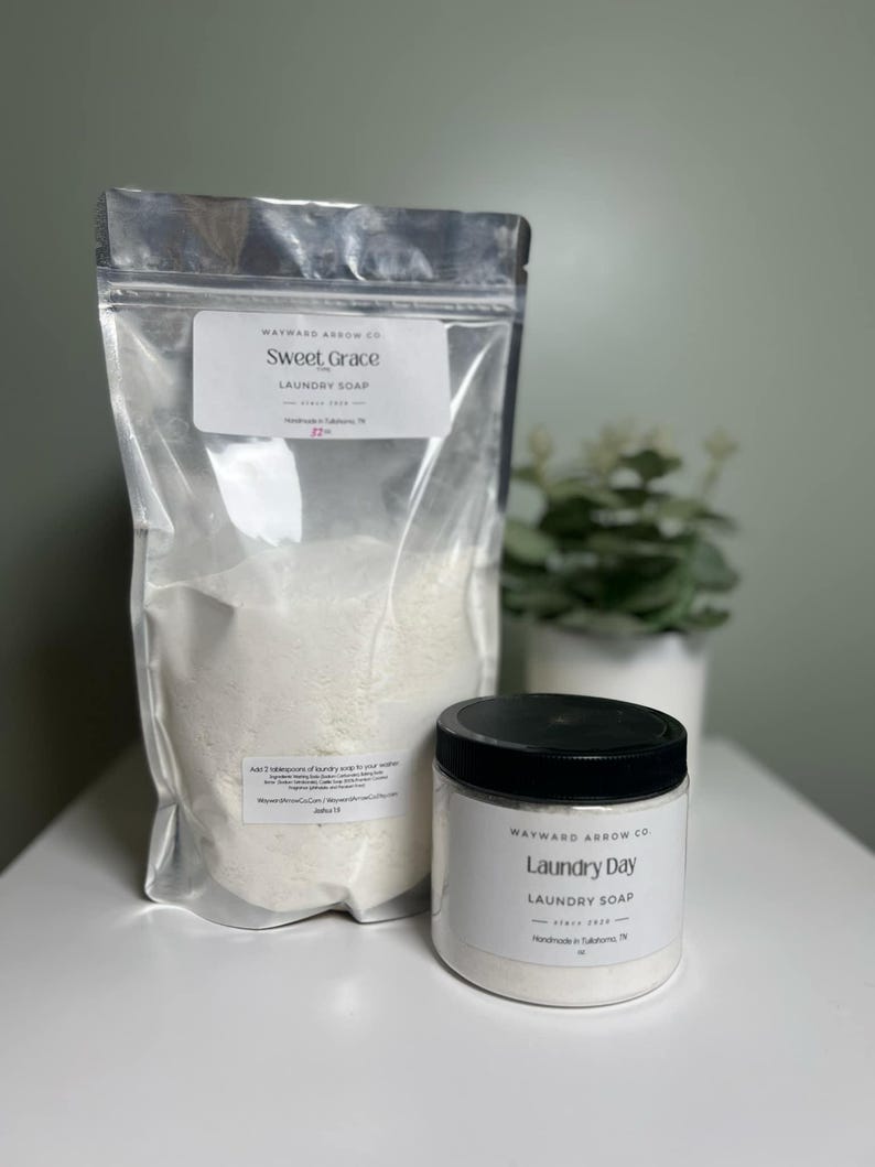May include: A clear plastic bag and a white jar of laundry soap sit on a white surface. The bag is labeled "Sweet Grace Laundry Soap" and the jar is labeled "Laundry Day Laundry Soap". A small plant in a white pot is in the background.