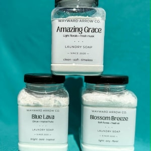 May include: Three clear jars of laundry soap with black lids. The jars are labeled "Amazing Grace," "Blue Lava," and "Blossom Breeze." The labels include the brand name, scent descriptions, and the phrase "LAUNDRY SOAP - SINCE 2020 -." The background is teal.