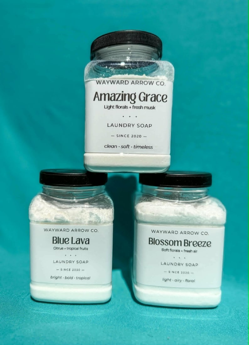 May include: Three clear jars of laundry soap with black lids. The jars are labeled "Amazing Grace," "Blue Lava," and "Blossom Breeze." The labels include the brand name, scent descriptions, and the phrase "LAUNDRY SOAP - SINCE 2020 -." The background is teal.