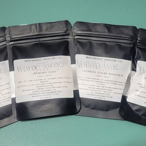May include: Four black pouches of laundry products, each with a white label. The labels read "Vanilla Luxe Laundry Soap", "Mango Tango Laundry Soap", "Vanilla Luxe Laundry Scent Booster", and "Wild West Laundry Scent Booster".