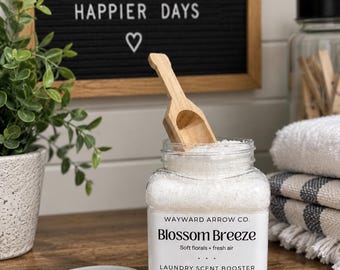 May include: A clear plastic jar filled with white laundry scent booster crystals, labelled Wayward Arrow Co. Blossom Breeze, with a wooden scoop inserted. The white lid sits beside the jar on a wooden surface, with a black letter board in the background.