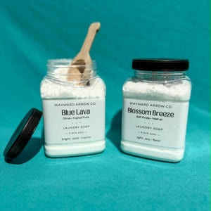 May include: Two clear jars of laundry soap, one with a wooden scoop. The jars have black lids and white labels with text: "Blue Lava" and "Blossom Breeze". The labels also read "WAYWARD ARROW CO." and "LAUNDRY SOAP".