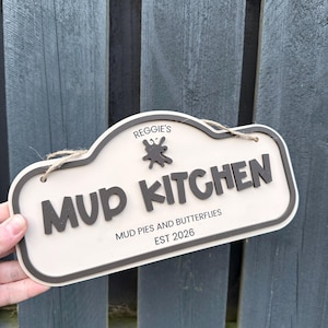 May include: A rectangular sign with rounded edges featuring a beige background and dark brown raised lettering that reads Reggie's Mud Kitchen, Mud Pies and Butterflies, and Est 2026, with a small dark brown mud splatter icon and a jute twine hanger.