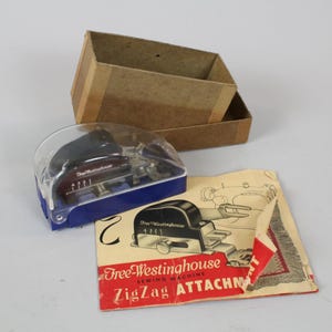 May include: An antique Free-Westinghouse Zig Zag sewing machine attachment, with original packaging. The attachment is in a clear plastic case with a blue base. Includes a cardboard box and a printed instruction sheet.