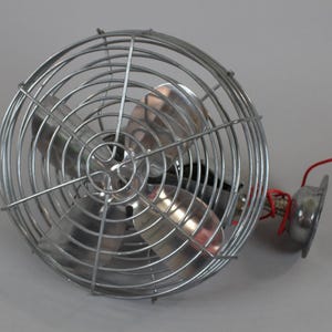 May include: A small, silver-colored electric fan with a protective wire cage. The fan has three blades and a red wire extending from the base. The fan is designed for personal use and is likely portable.