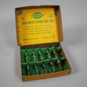 Op de afbeelding: Een gele doos met groene rand die een set Greenlee solid-center elektrische boorbits bevat. De doos is gelabeld "Set Solid-Center Electric Drill Bits" en geeft de maten van de bits weer: 4/16", 3/16", 1/4", 5/16", 3/8", 7/16", 1/2", 5/8", en 3/4". De doos vermeldt ook dat de bits een 3-inch draailengte hebben en zijn ontworpen om te passen op 1/4-inch elektrische boormachines. De doos is gelabeld "Greenlee Tool Co. Rockford, Ill. U.S.A."