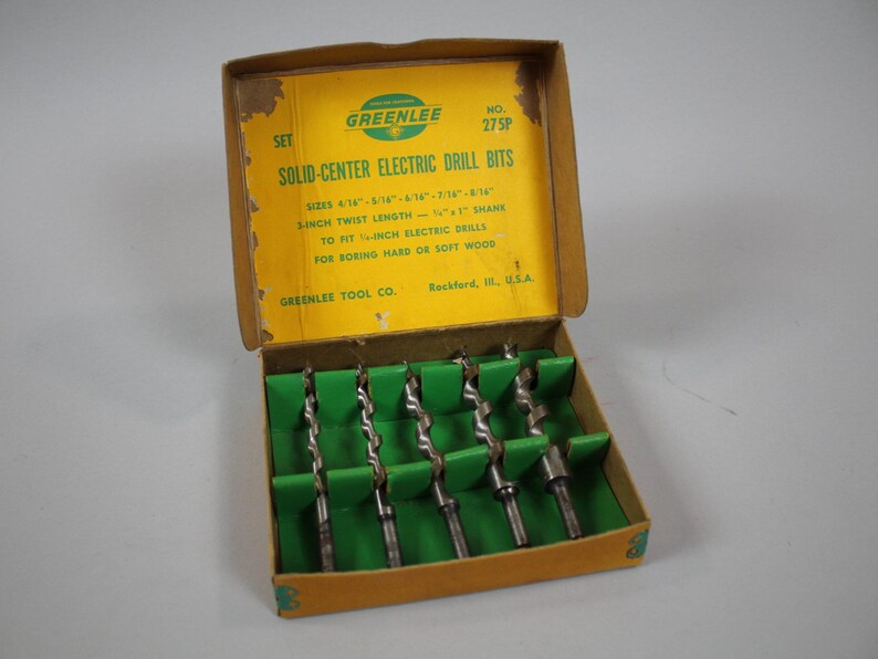Op de afbeelding: Een gele doos met groene rand die een set Greenlee solid-center elektrische boorbits bevat. De doos is gelabeld "Set Solid-Center Electric Drill Bits" en geeft de maten van de bits weer: 4/16", 3/16", 1/4", 5/16", 3/8", 7/16", 1/2", 5/8", en 3/4". De doos vermeldt ook dat de bits een 3-inch draailengte hebben en zijn ontworpen om te passen op 1/4-inch elektrische boormachines. De doos is gelabeld "Greenlee Tool Co. Rockford, Ill. U.S.A."