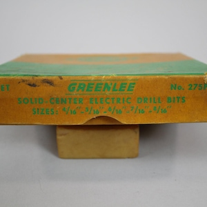 Op de afbeelding: Een bruine kartonnen doos met groene rand en de tekst "GREENLEE" in groene letters. De doos is gelabeld "SET SOLID-CENTER ELECTRIC DRILL BITS" en vermeldt de maten van de boortjes: "4/16 - 5/16 - 6/16 - 7/16 - 8/16". De doos is gelabeld "No. 275P".