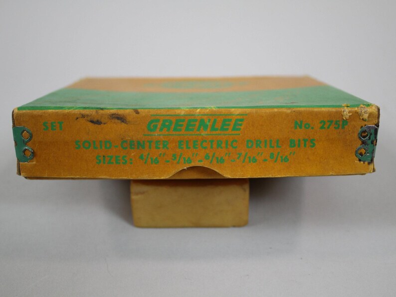 Op de afbeelding: Een bruine kartonnen doos met groene rand en de tekst "GREENLEE" in groene letters. De doos is gelabeld "SET SOLID-CENTER ELECTRIC DRILL BITS" en vermeldt de maten van de boortjes: "4/16 - 5/16 - 6/16 - 7/16 - 8/16". De doos is gelabeld "No. 275P".