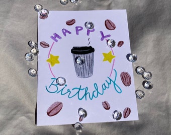 May include: A white card with the words "Happy Birthday" written in purple and teal. A cartoon coffee cup with steam coming out of it is in the center of the card. There are coffee beans and stars around the coffee cup.