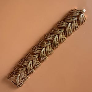 May include: A gold-tone metal bracelet featuring a repeating pattern of textured, fan-shaped leaf or feather motifs. The links are arranged in a dense, overlapping design that creates a continuous, articulated band with a metallic finish.
