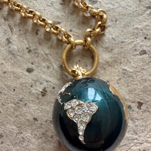 May include: A gold-coloured necklace with a globe pendant. The globe is dark teal with gold detailing and a crystal design representing South America. The chain is a chunky link style.