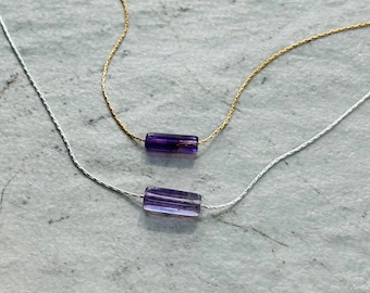 May include: Two delicate necklaces with purple gemstone pendants. The necklaces are on a white textured background. The top necklace has a gold chain and the bottom necklace has a silver chain.