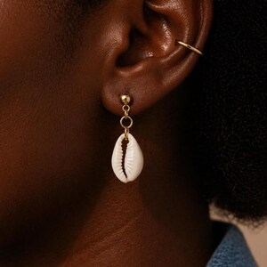 May include: A gold-colored earring with a small, round stud and a dangling white seashell. The earring is attached to the ear with a small gold hoop. The seashell has a natural, textured appearance.