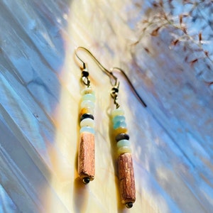 Peut inclure: Boucles d'oreilles pendantes avec crochets dor&eacute;s. Chaque boucle d'oreille pr&eacute;sente une rang&eacute;e verticale de petites perles color&eacute;es, dont du bleu clair, du jaune, du noir et du marron, au-dessus d'un cylindre en bois marron. Pr&eacute;sent&eacute;es sur une surface nacr&eacute;e.