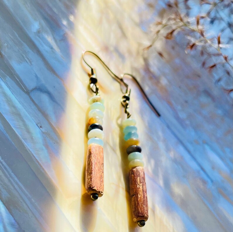 Peut inclure: Boucles d'oreilles pendantes avec crochets dor&eacute;s. Chaque boucle d'oreille pr&eacute;sente une rang&eacute;e verticale de petites perles color&eacute;es, dont du bleu clair, du jaune, du noir et du marron, au-dessus d'un cylindre en bois marron. Pr&eacute;sent&eacute;es sur une surface nacr&eacute;e.