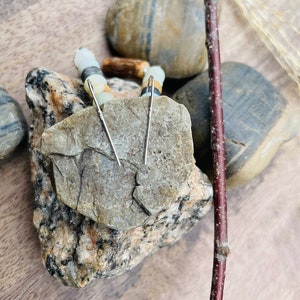 Peut inclure: Une paire de boucles d'oreilles en argent en forme de barres, pr&eacute;sent&eacute;es sur une roche grise textur&eacute;e. Les boucles d'oreilles ont un design simple et lin&eacute;aire. L'arri&egrave;re-plan comprend des pierres lisses et multicolores et une brindille brun fonc&eacute;, cr&eacute;ant une esth&eacute;tique naturelle et terreuse.