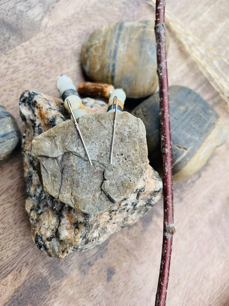 Peut inclure: Une paire de boucles d'oreilles en argent en forme de barres, pr&eacute;sent&eacute;es sur une roche grise textur&eacute;e. Les boucles d'oreilles ont un design simple et lin&eacute;aire. L'arri&egrave;re-plan comprend des pierres lisses et multicolores et une brindille brun fonc&eacute;, cr&eacute;ant une esth&eacute;tique naturelle et terreuse.