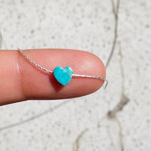 May include: A small, turquoise heart-shaped bead strung on a delicate silver chain. The bead is held between a finger and thumb, showcasing its vibrant color and intricate detail. The chain appears to be approximately 0.5 mm in diameter.