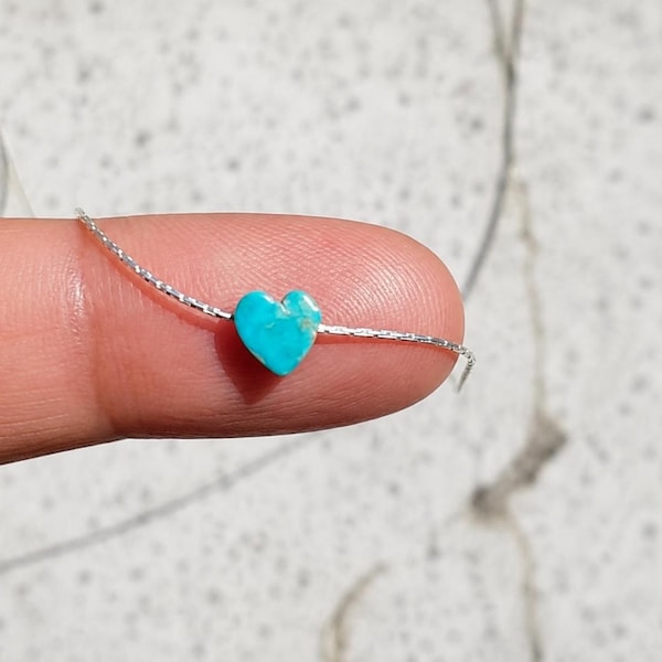 May include: A small, turquoise heart-shaped bead strung on a delicate silver chain. The bead is held between a finger and thumb, showcasing its vibrant color and intricate detail. The chain appears to be approximately 0.5 mm in diameter.