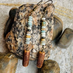 Peut inclure: Boucles d'oreilles faites &agrave; la main avec une cha&icirc;ne verticale de petites perles color&eacute;es dans les tons de bleu, blanc, beige et marron. Les perles sont reli&eacute;es par un fil de couleur laiton et surmont&eacute;es d'un charm cylindrique en bois. Les boucles d'oreilles sont suspendues &agrave; des crochets d'oreilles couleur laiton.