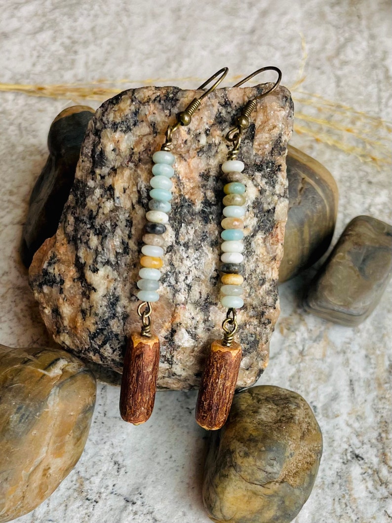 Peut inclure: Boucles d'oreilles faites &agrave; la main avec une cha&icirc;ne verticale de petites perles color&eacute;es dans les tons de bleu, blanc, beige et marron. Les perles sont reli&eacute;es par un fil de couleur laiton et surmont&eacute;es d'un charm cylindrique en bois. Les boucles d'oreilles sont suspendues &agrave; des crochets d'oreilles couleur laiton.
