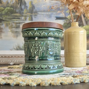 Può includere: Un vaso di ceramica verde con un coperchio di legno e intricati disegni in rilievo. Il vaso è appoggiato su un sottopentola di pizzo accanto a un vaso di ceramica giallo.