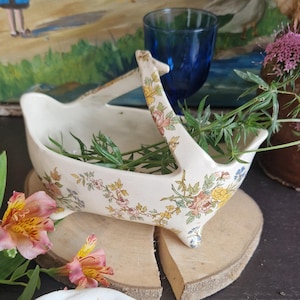 Puede incluir: Una cesta de cerámica crema con asa y estampados florales, apoyada sobre una rodaja de madera. La cesta está llena de ramitas verdes. Flores rosas y amarillas y un lirio de agua blanco están en primer plano. Un vaso azul y una pintura están en el fondo.