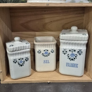 May include: A wooden shelf displays three ceramic canisters with lids, and a larger bowl. The canisters are white with blue floral designs and the words "SEL" and "FARINE". The bowl and canisters are vintage kitchenware.
