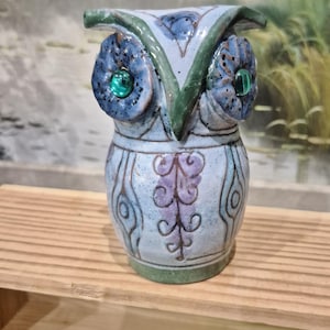 May include: Ceramic owl figurine with blue, green, and purple accents. The owl has large blue eyes with green centers, and a decorative pattern on the body. The figurine is on a wooden shelf.