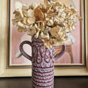 May include: A red and blue ceramic vase with two handles, filled with dried beige flowers. The vase has a textured, patterned design. A framed painting of a girl in a pink dress is in the background.