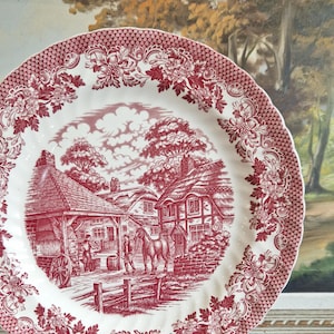 Könnte beinhalten: Ein vintage Teller aus roter und weißer Keramik mit einer detaillierten Illustration einer ländlichen Szene mit Häusern, einem Pferd und einem Wagen. Der Teller hat einen gewellten Rand und ein Blumenmuster.