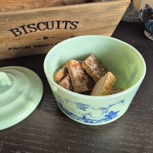 Puede incluir: Un tarro de galletas de cerámica verde claro con tapa, lleno de varias galletas. El tarro tiene un diseño decorativo azul y blanco. Una caja de madera con la palabra "BISCUITS" está en el fondo.