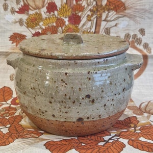 Może przedstawiać: Ceramiczny garnek z pokrywką, pokryty pstrokatą glazurą w odcieniach brązu, beżu i bieli. Garnek ma zaokrąglone dno i dwie rączki po bokach.